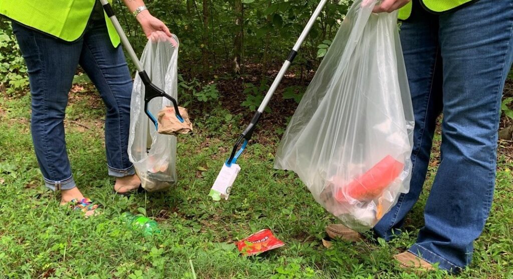 Photograph of two people from the waist down that are using grabbers to pick up trash from the ground and put it in a trash bag